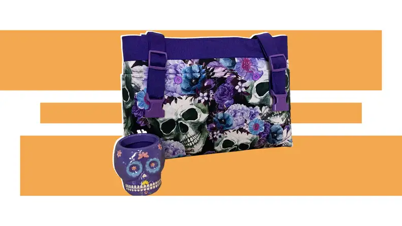 A purple bag with a skull and floral design sits next to a purple calavra mug from moBellaBags in front of an orange and white design.