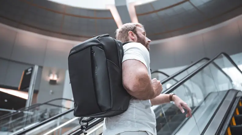 A man stands ascends on an escalator at the airport, wearing a Briggs and Riley Cargo Backpack