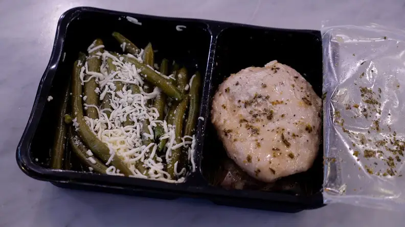 Apricot-glazed chicken and cheese-covered green beans inside of black container on countertop.