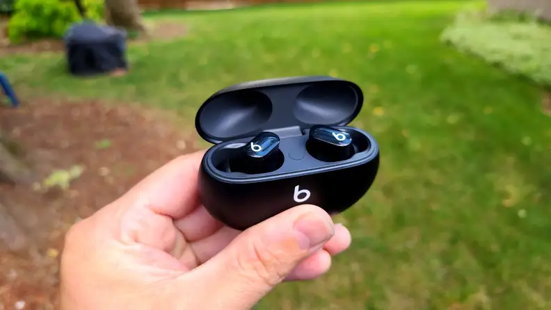 The black Beats Studio Buds are held with the case open in front of a lush lawn and red mulch.