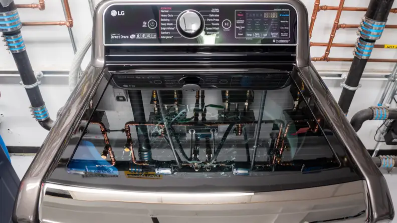 A close-up of an LG top-loader in our testing labs. Behind the appliance and in the reflection on its translucent lid you can see a network of pipes we use to regulate water temperature and flow.