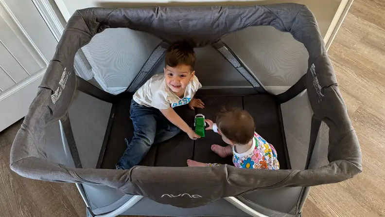 Two small toddlers sit inside of the Nuna Sena Aire while looking up at the camera.