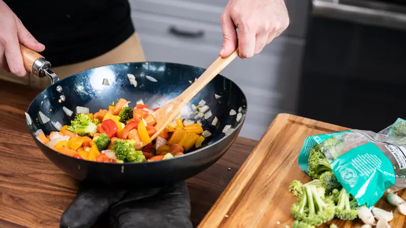 A person is using a spatula to stir a medley of vegetables in a carbon steel wok.
