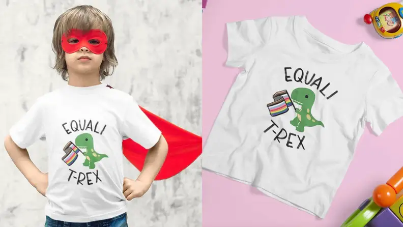 Child wearing a red cape posing like a superhero wearing a white tshirt with a dinosaur on it holding a rainbow flag with the pun "Equali T-Rex". Next to that, is the same white tshirt lying on a pink background.