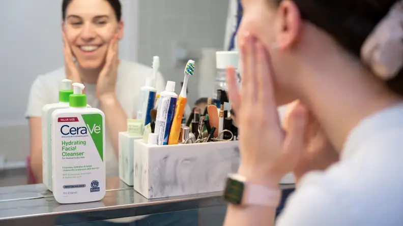 In a mirror you can see a person's reflection as they wash their face and a bottle of face wash sits in front of them on a shelf.