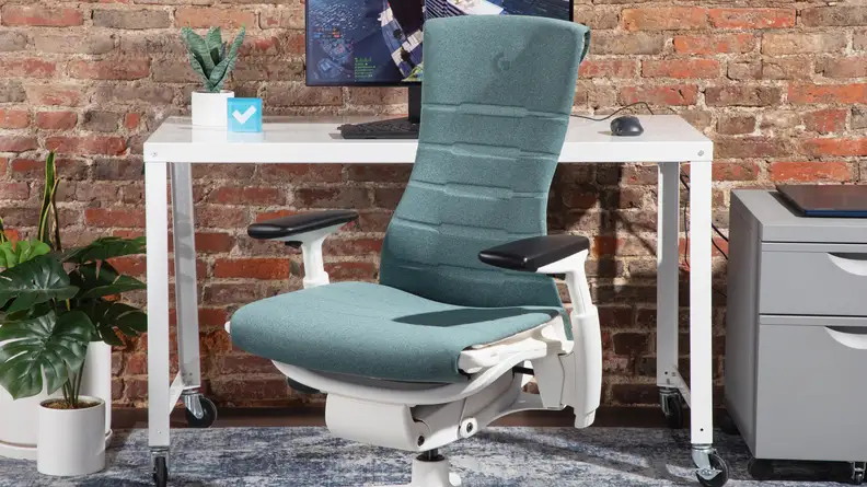The Herman Miller Embody gaming chair in the color blue, next to a white office desk and office supplies.
