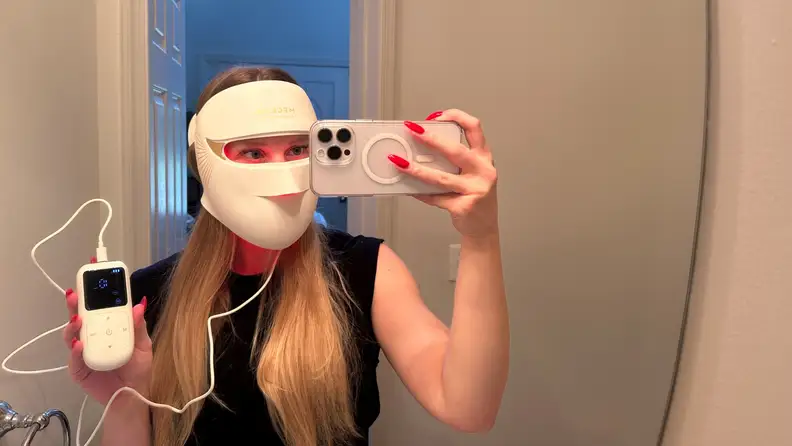 Blonde woman wearing the Megelin LASER & LED mask, holding the remote in front of a mirror.