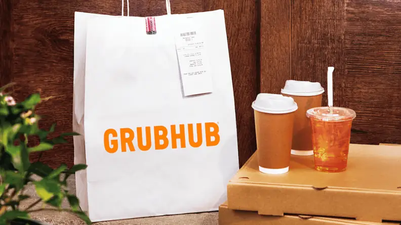 White GrubHub bag next to two coffee cups and an iced tea in front of a dark wooden background.