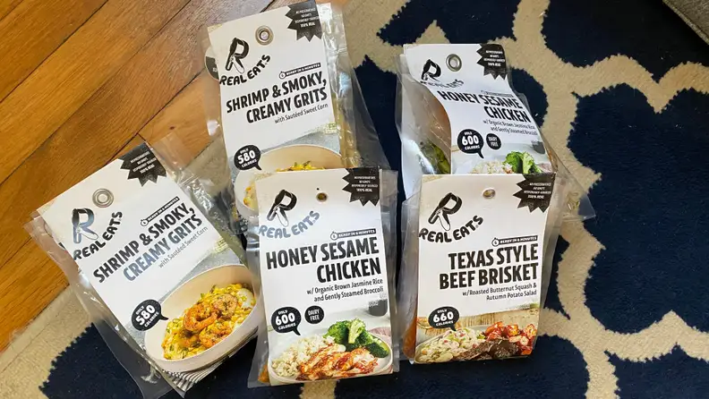 Five packages of RealEats premade meals are in the picture. From the left: Shrimp with creamy grits, honey sesame chicken, and Texas style beef brisket.