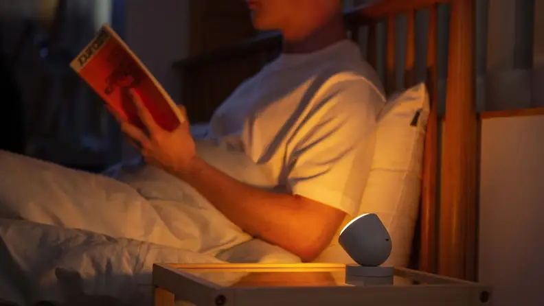 A person reads a book in bed with the help of an orange-hued Olly Night light.