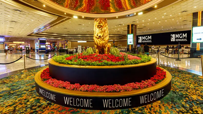 MGM Grand hotel lobby featuring the resort’s signature golden lion statue surrounded by floral displays, with check-in counters and guests in the background.