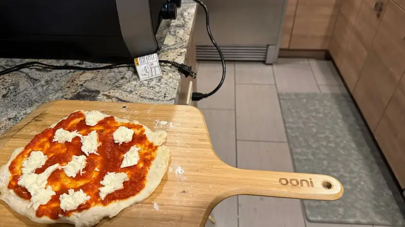 Raw pizza pie sitting on top of wooden pizza peel next to the Ooni Volt pizza oven.