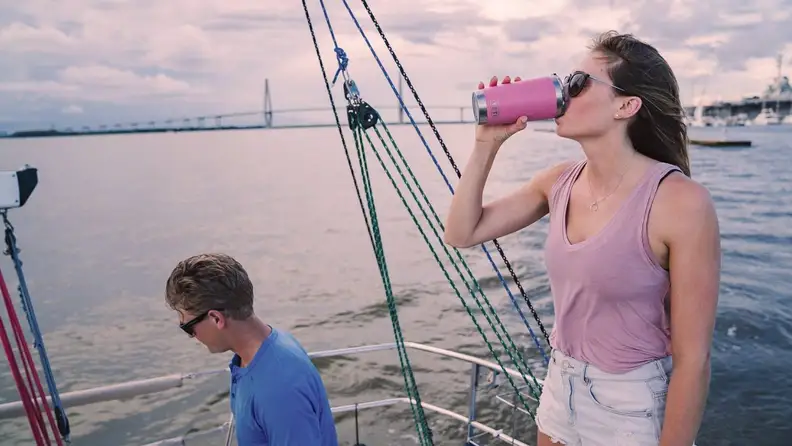 Two people on a boat, the woman is standing up and drinking from a reusable mug