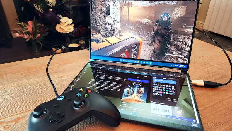 A dual-screen laptop showing a videogame on one display and a website on the other. An Xbox controller is connected to the laptop with a cable.