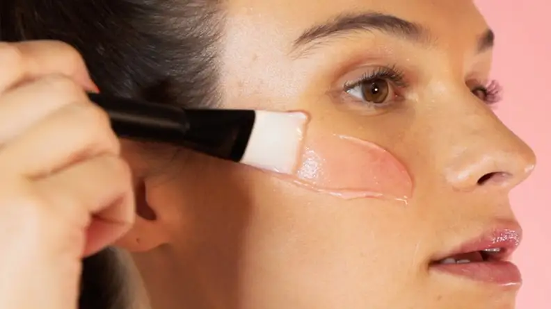 The profile of a model's face as she uses a brush to apply a pink liquid to her face.
