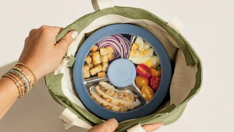 Divided stainless steel lunch container filled with chicken, eggs, vegetables, and croutons inside a reusable tote bag.