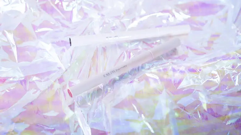 A gray tube of lip stain marker front and center with a white tube of liquid eyeliner in the background, both on holographic cellophane paper.