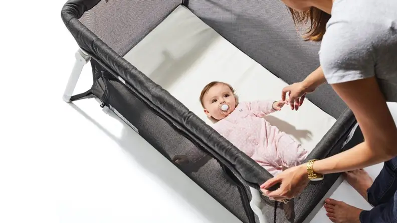 Baby in pink pajamas with a pacifier in their mouth looking up from a bassinet to their caregiver.