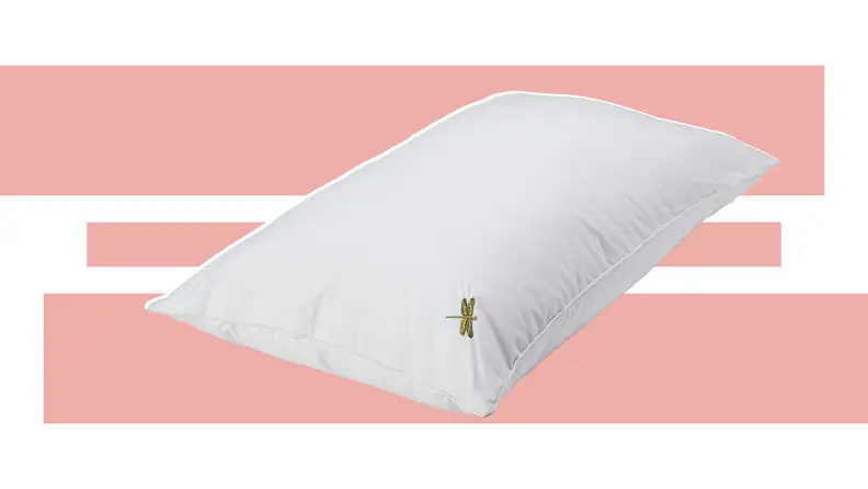 A white DreamPad pillow with a small dragonfly design embroidered in the corner in front of a pink and white background.