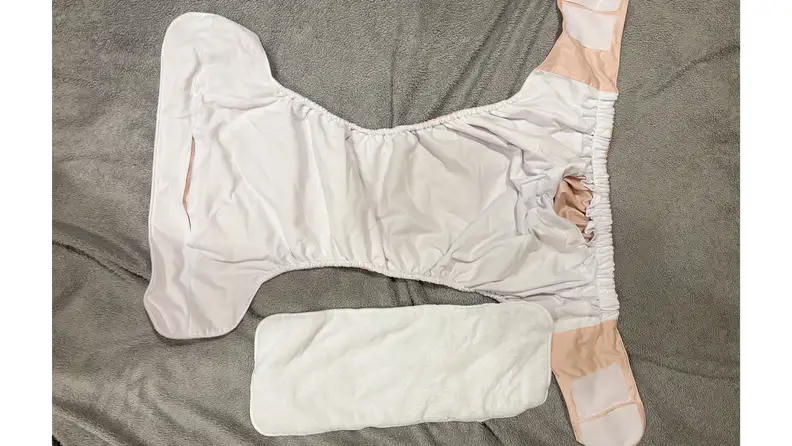 A peach diaper with hook-and-loop closures laid open next to an insert on a gray fabric surface.