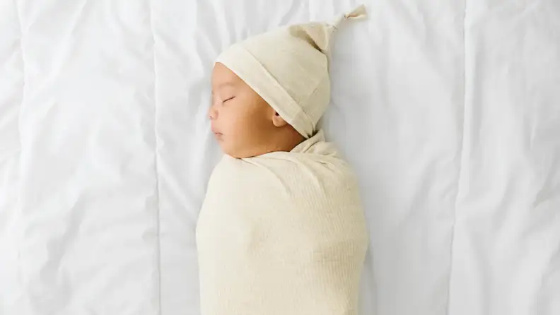 Swaddled baby peacefully sleeping on a white bed, wearing a cream-colored hat and wrap.