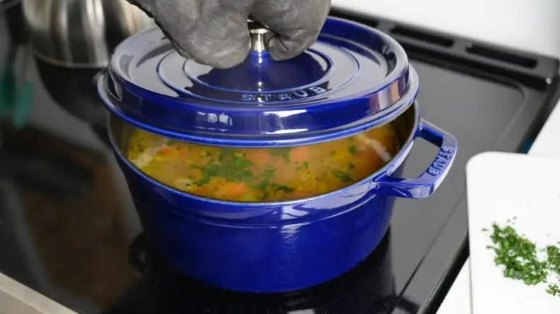 A chef lifting the lid on a Staub Dutch oven, revealing soup inside.