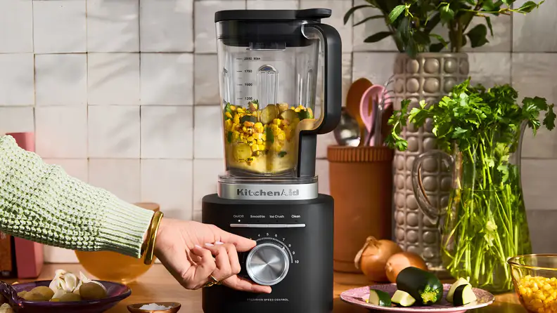Person adjusting the speed dial on a KitchenAid blender filled with vegetables and ingredients on a kitchen countertop.