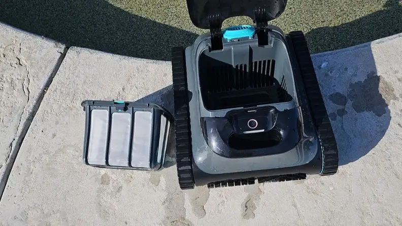 The Aiper Scuba S1 and its collection basket sits closely to the edge of a pool on a sunny day outdoors.