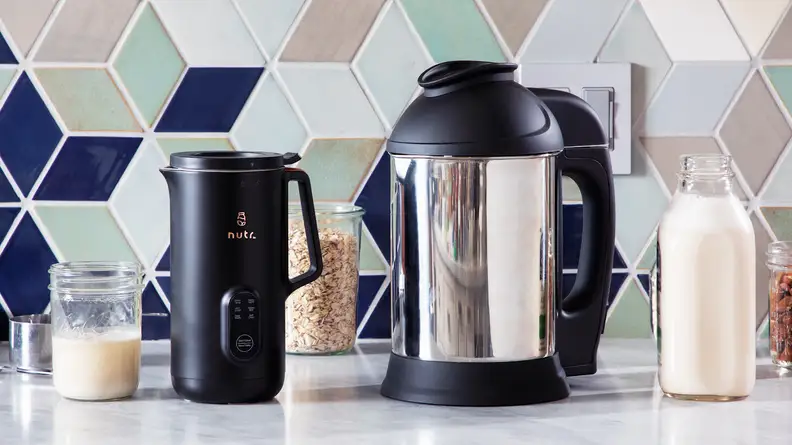 The Nutr plant-based milk maker next to the Almond Cow plant-based milk maker on granite countertop in front of tiled wall and 3 clear jars with snacks inside and one glass jar of milk.