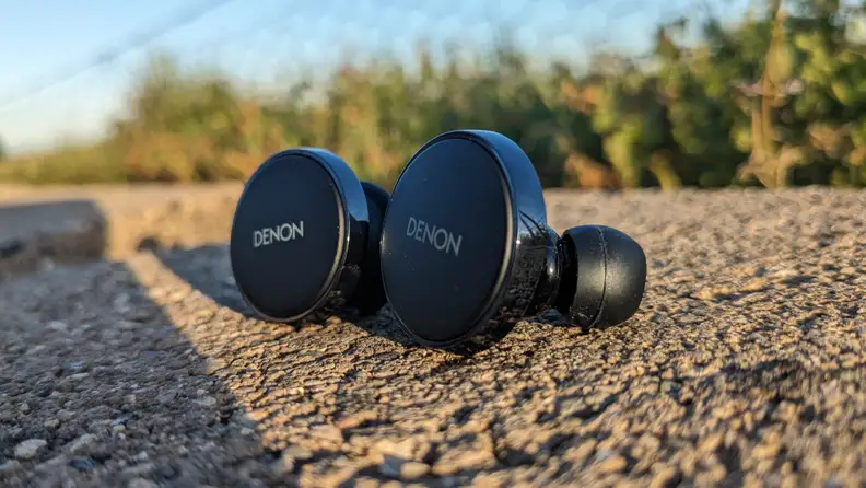A side view of a pair of Denon PerL Pro earbuds on top of concrete on a sunny day outdoors.