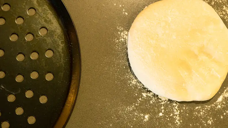 A Made In carbon steel perforated pizza pan is next to a pizza dough.