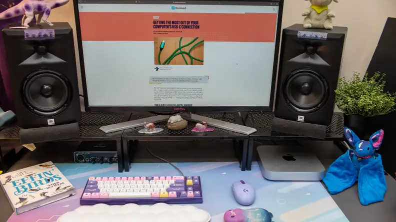 Desktop set-up featuring the Apple Mac Mini M2, a desktop computer, two speakers, a colorful computer keyboard, a plush stuffed animal and a book.