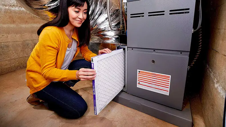 A woman kneels beside a furnace, changing in a new Filtrete Micro Allergen Defense Air Filter.
