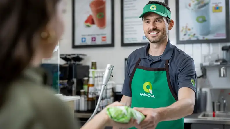 A smiling QuickChek employee handing a bag to a customer at the counter