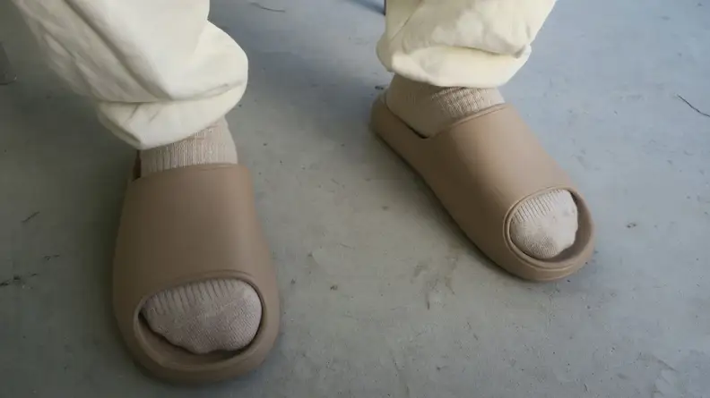 A pair of feet standing in beige pool slide sandals from Litfun, in off-white socks and cream colored sweatpants.