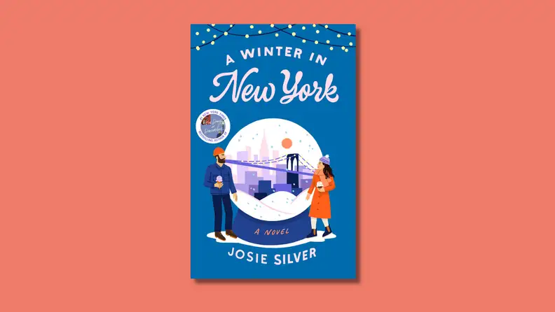 An image of the cover of "A Winter in New York," by Josie Silver, featuring two people in wintery clothing in front of a snow globe containing a vignette of New York City.