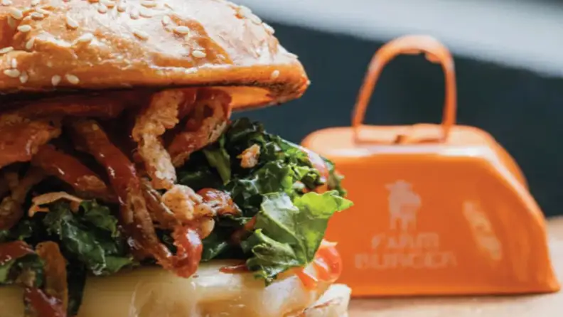 Close-up of a loaded burger with crispy onions and greens, next to a Farm Burger bell