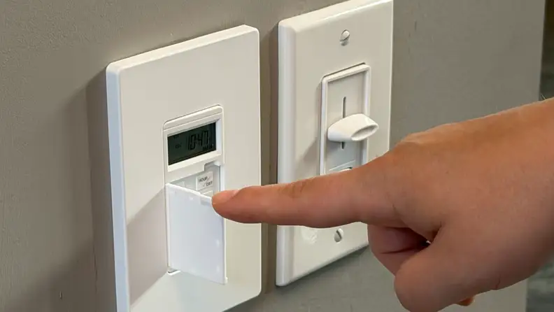 White Lider 7-Day Programmable Digital Timer Switch shown next to a dimmer switch and a hand pressing the buttons on a grey wall.