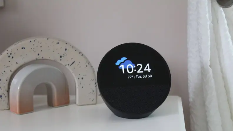 The Amazon Echo Spot on a nightstand, displaying the time and weather next to a small marble statue.