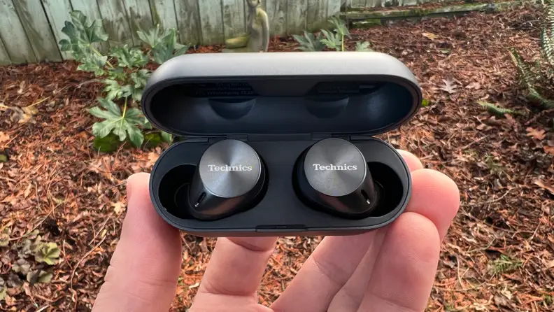 The gunsmoke colored wireless earbuds sit in their charging case with the lid open and brown debris in the background.n