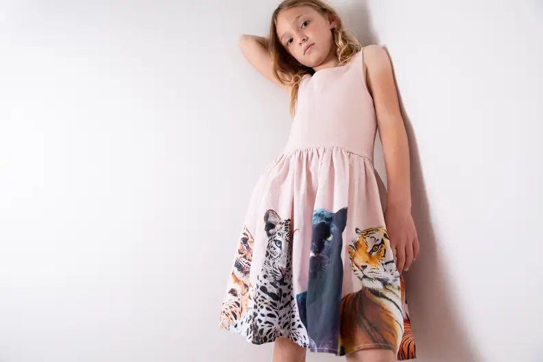 A girl wearing a pink dress printed with cats leans against a wall.