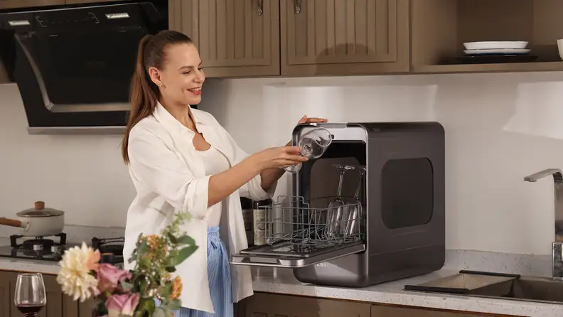 A woman loads wine glasses into the Lissome R1 compact countertop dishwasher on a kitchen counter.