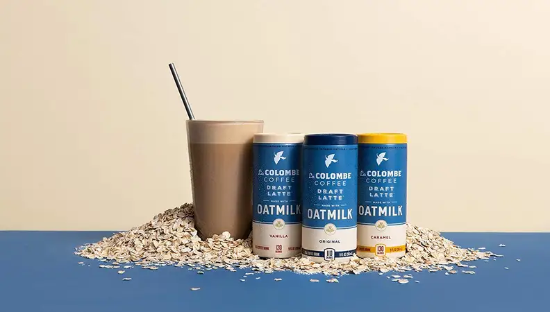 A grouping of canned oat milk lattes sits on a bed of oats accompanied by a latte in a glass.