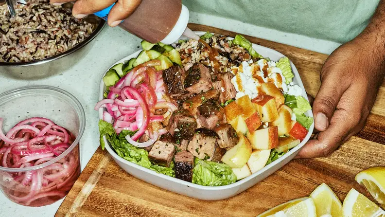 A hand drizzling dressing over a salad bowl with steak, apples, and grains