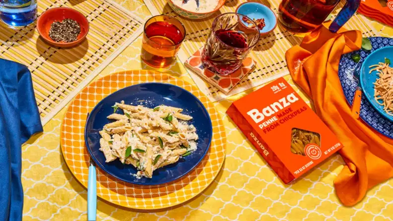 Box of Banza chickpea penne beside a plated pasta dish and drinks