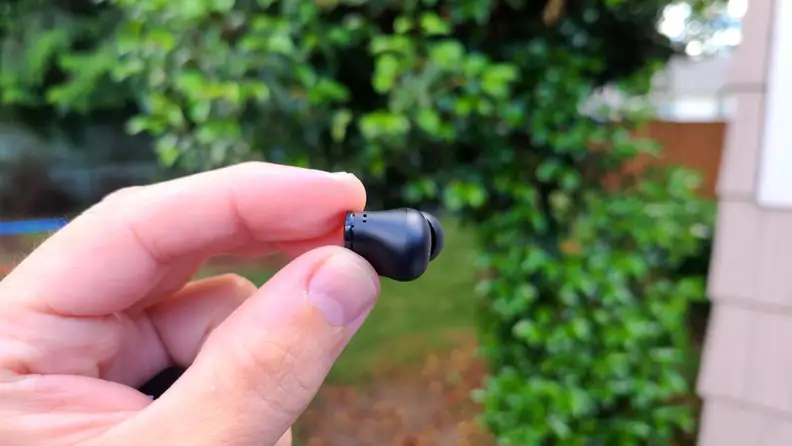 The black Beats Studio bud is held in fingers in front of a green bush with a beige house at the right.