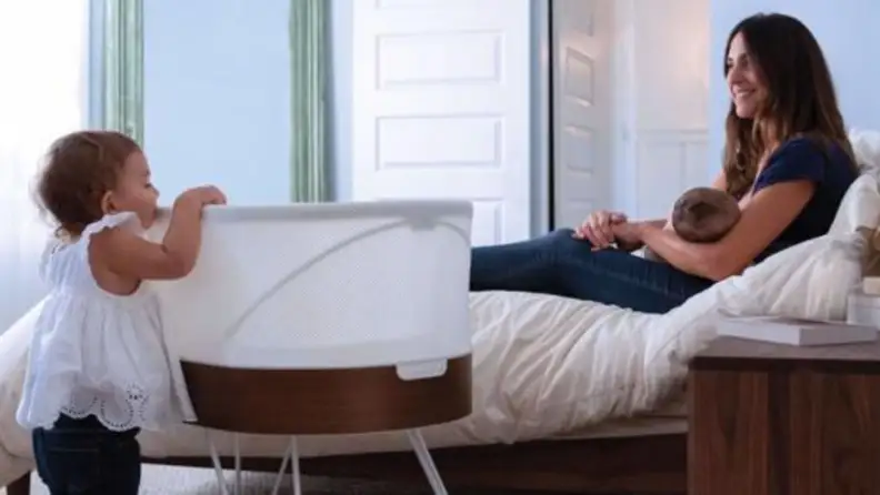toddler looking into the SNOO bassinet while mother smiles on holding baby.