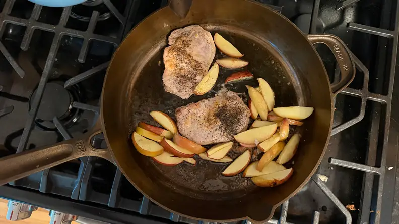 A cast iron pan on a stove with cooked pork and apples