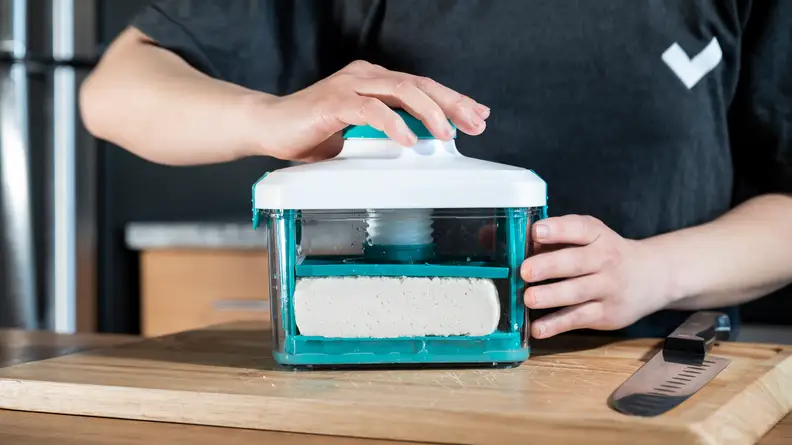 A person using the Noya Tofu Press to press a block of tofu on a wooden cutting board.