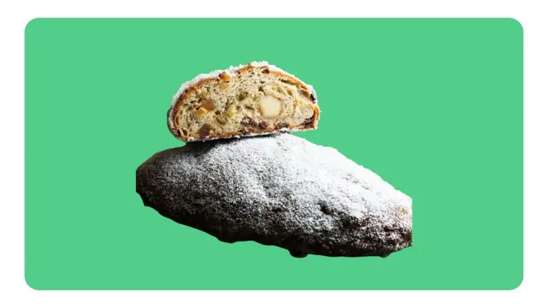 A whole powdered German stollen loaf next to one sliced in half in front of green background.